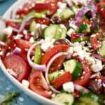 A close-up of tomato wedges, onion, cucumber and bell pepper slices topped with a light vinaigrette and feta cheese in a large white serving platter.
