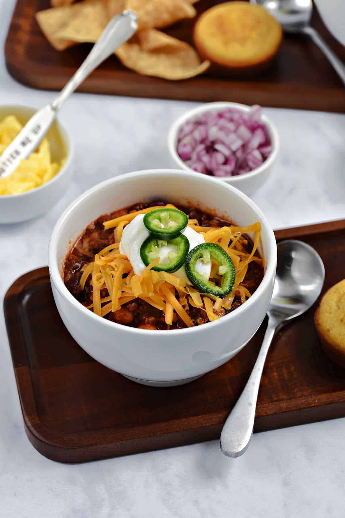 A bowl of chili topped with sour cream, cheddar cheese, and sliced jalapeños on a board with cornbread muffin and tortilla chips.