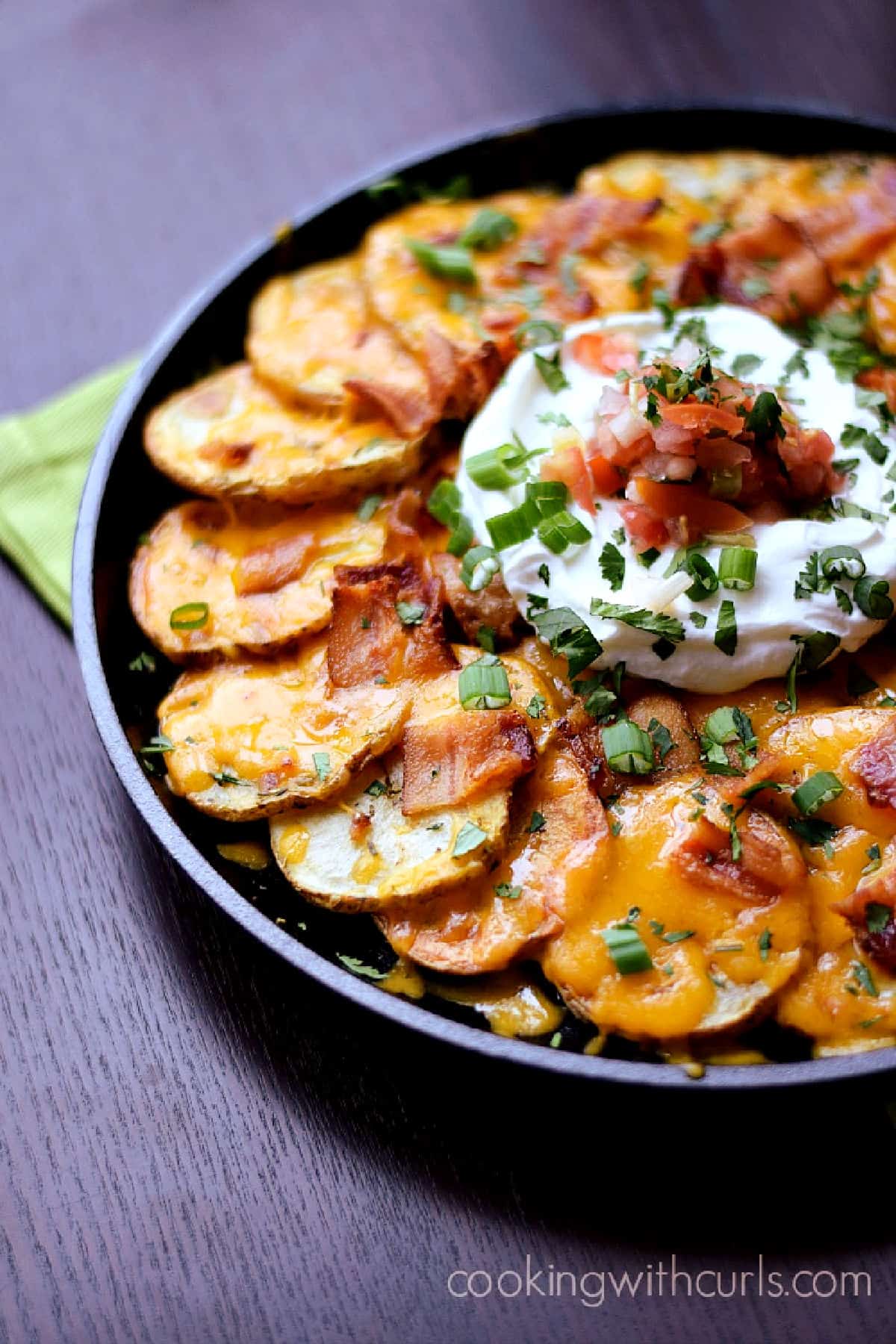 Thinly sliced potatoes arranged in a cast iron skillet topped with melted cheese, bacon, sour cream, salsa and green onions.