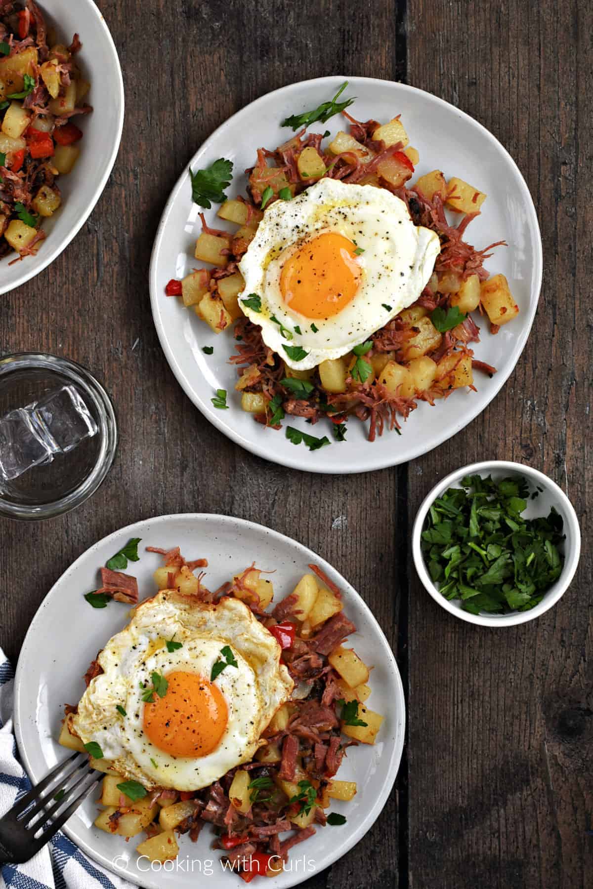Looking down on two plates with corned beef hash topped with a fried egg and chopped parsley.