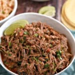 A bowl of Mexican shredded beef surrounded by bowls of rice and black beans, with corn tortillas in the background.
