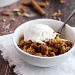 A white bowl with apple crisp topped with vanilla ice cream sitting on a white fringed napkin with an apple and baking dish in the background.