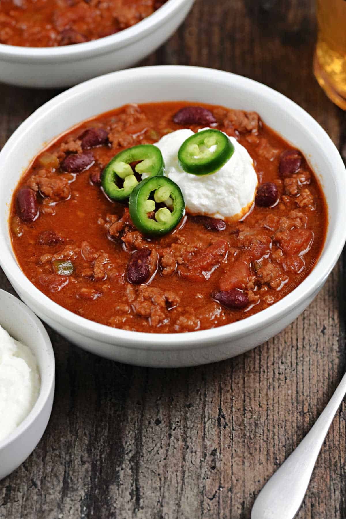 A bowl of chili topped with sour cream and sliced jalapeños with a small bowl of sour cream on the side.