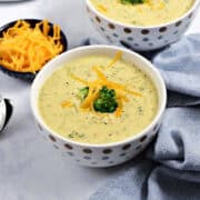 Two bowls of broccoli cheddar soup topped with broccoli and grated cheddar cheese.